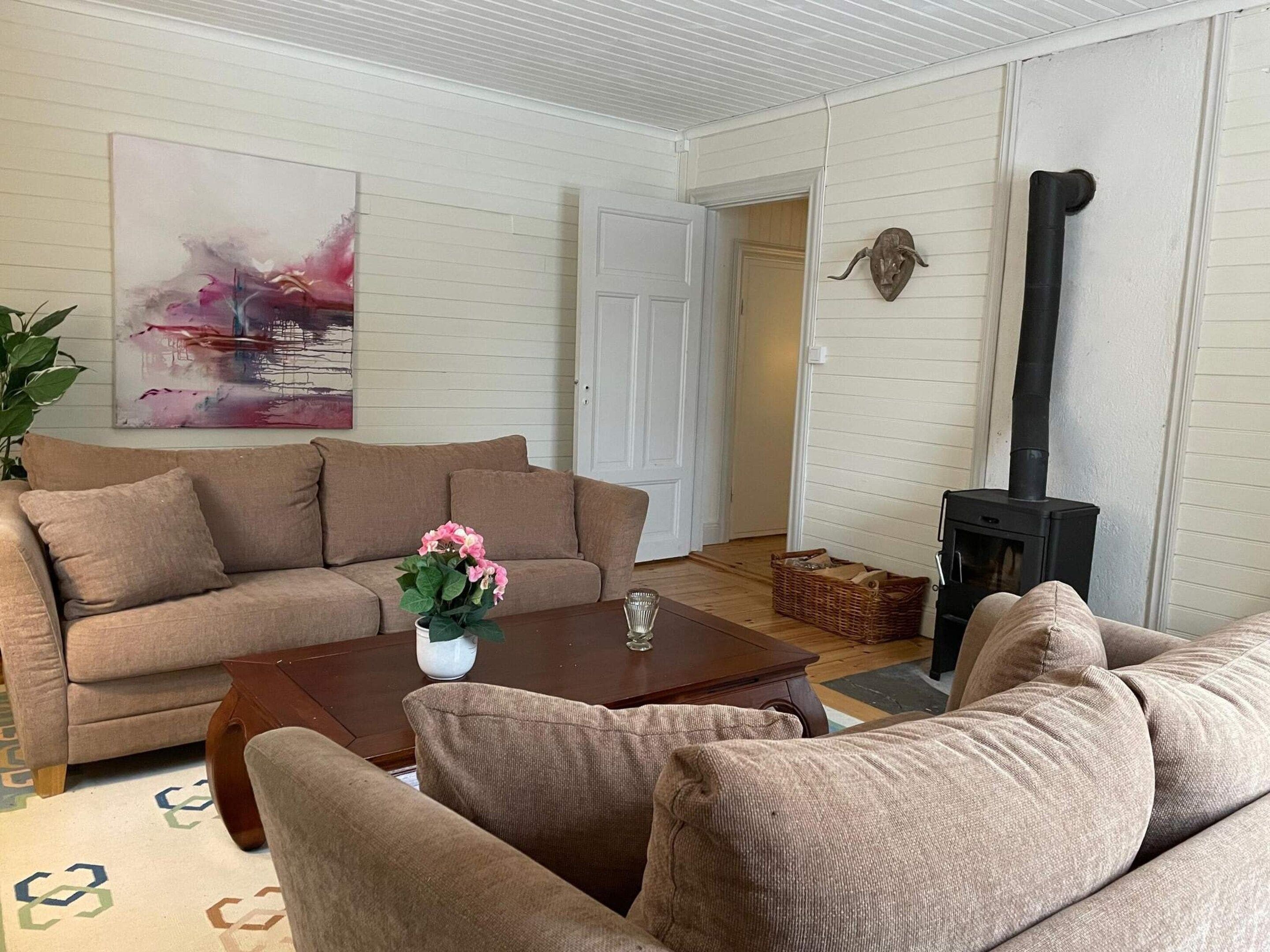 A living room with a beige sofa, a coffee table, a vase with flowers, a painting on the wall, and a wood-burning stove.