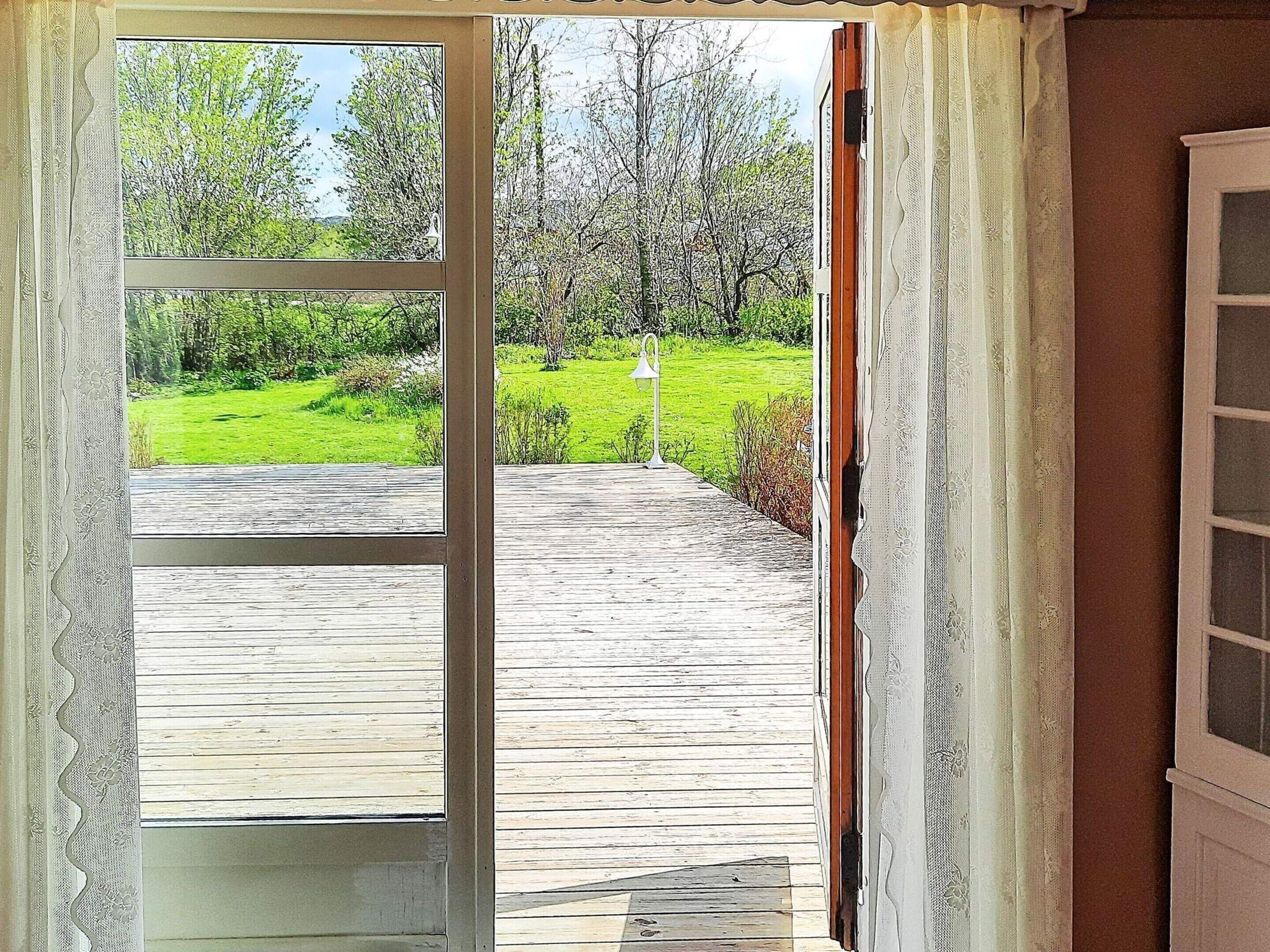 Una puerta abierta que da a una terraza de madera, con un área de césped y árboles al fondo.