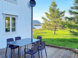 A deck with a glass table and chairs, overlooking a grassy area with trees and a body of water.