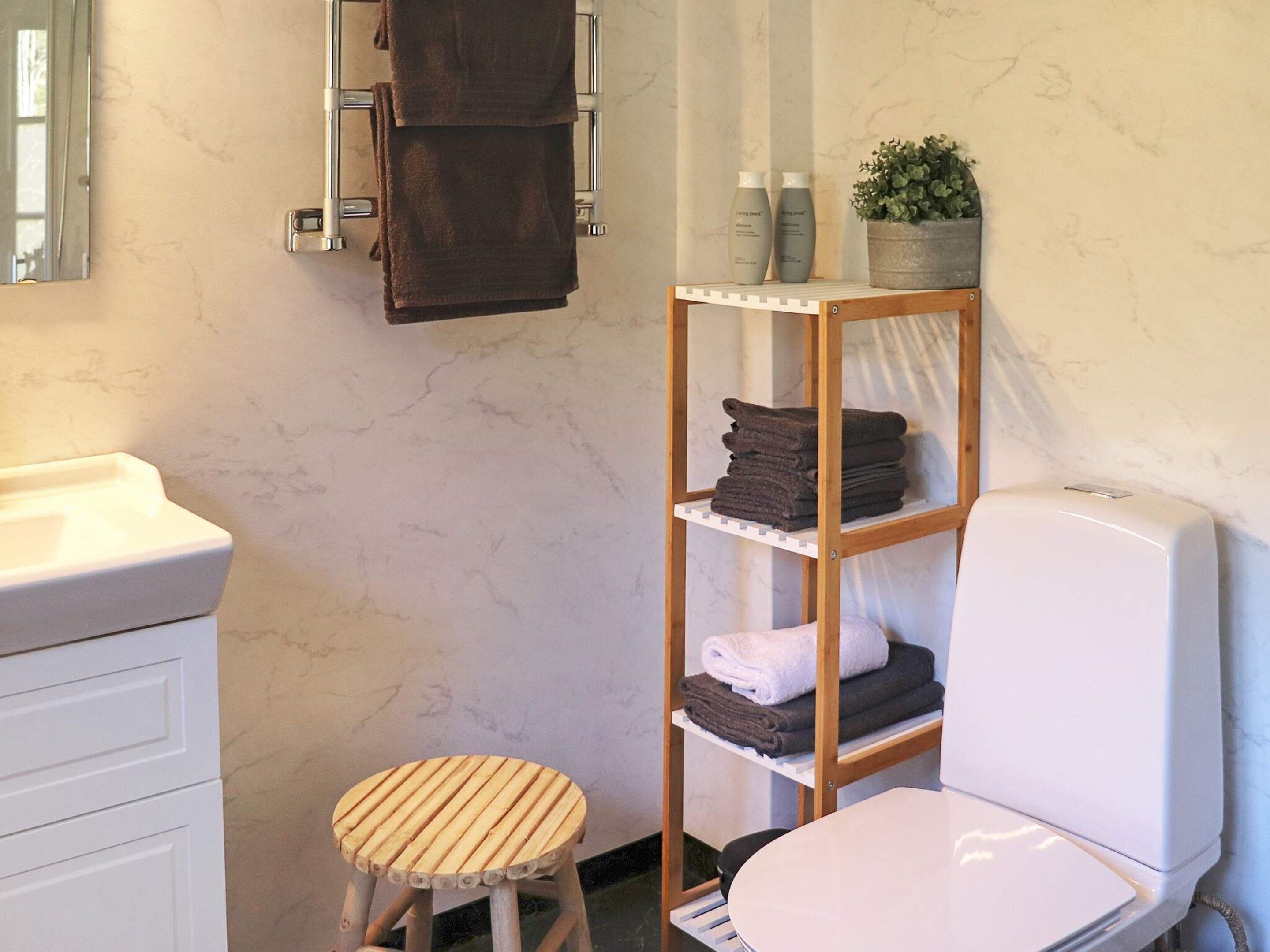 Une salle de bain comprenant des toilettes, un lavabo, un miroir, un porte-serviettes, une étagère avec des serviettes et un petit tabouret.