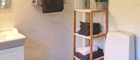 A bathroom with a toilet, a sink, a mirror, a towel rack, a shelf with towels, and a small stool.
