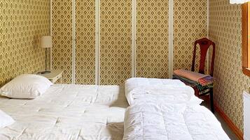 A room with two beds, a small table, a chair, and patterned wallpaper.