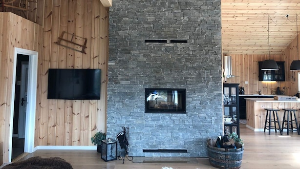 A modern interior with a stone fireplace, wooden walls, and a kitchen area. - 4 Star Holiday Home in Skare (Ullensvang)