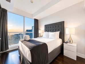 A hotel room with a large bed, a nightstand, a lamp, and a view of the city through large windows.