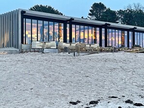 Moderni rakennus, jossa on suuret lasi-ikkunat, ulkotila istumiseen ja hiekkapohja.