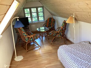 Een gezellige zolderkamer met een bed, twee rieten stoelen, een klein tafeltje, een televisie en een raam met uitzicht op bomen.