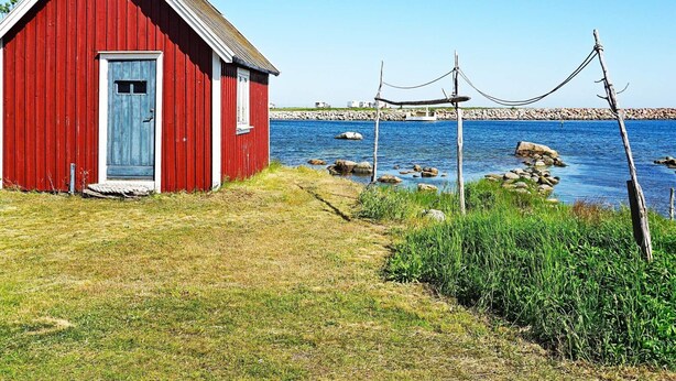 A red wooden building with a blue door, situated by a rocky shoreline with a clear blue sky. - 6 Person Holiday Home in Solvesborg-by Traum (Sölvesborg)