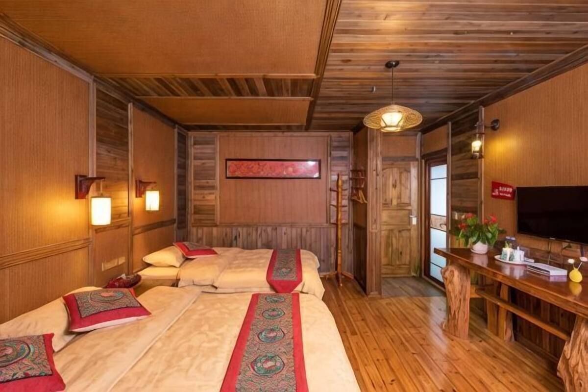 A room with wooden floors, a bed with a red and green patterned bedspread, a wooden bench, a wall-mounted TV, and a wooden door.