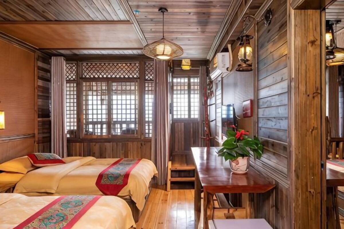A traditional room with wooden walls, a wooden ceiling, a window with a lattice design, a bed with a red and green patterned bedspread, and a table with a potted plant.