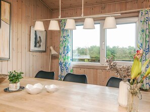 A wooden interior with a dining table, chairs, a lamp, and a window with curtains.