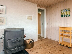 A room with a wood-burning stove, a metal trash bin, a wooden shelf unit, and a white brick wall with framed pictures. - 6 Person Holiday Home in Fano (Fano)