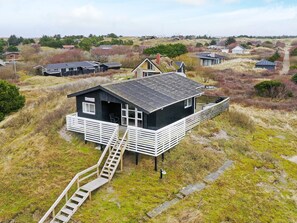 A black house with a white porch and a wooden staircase leading up to it, surrounded by grassy fields and other houses. - 6 Person Holiday Home in Fano (Fano)