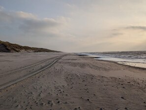 A sandy beach with tire tracks leading to the water, a dune on the left, and a cloudy sky. - 12 Person Holiday Home in Ulfborg (Ulfborg)