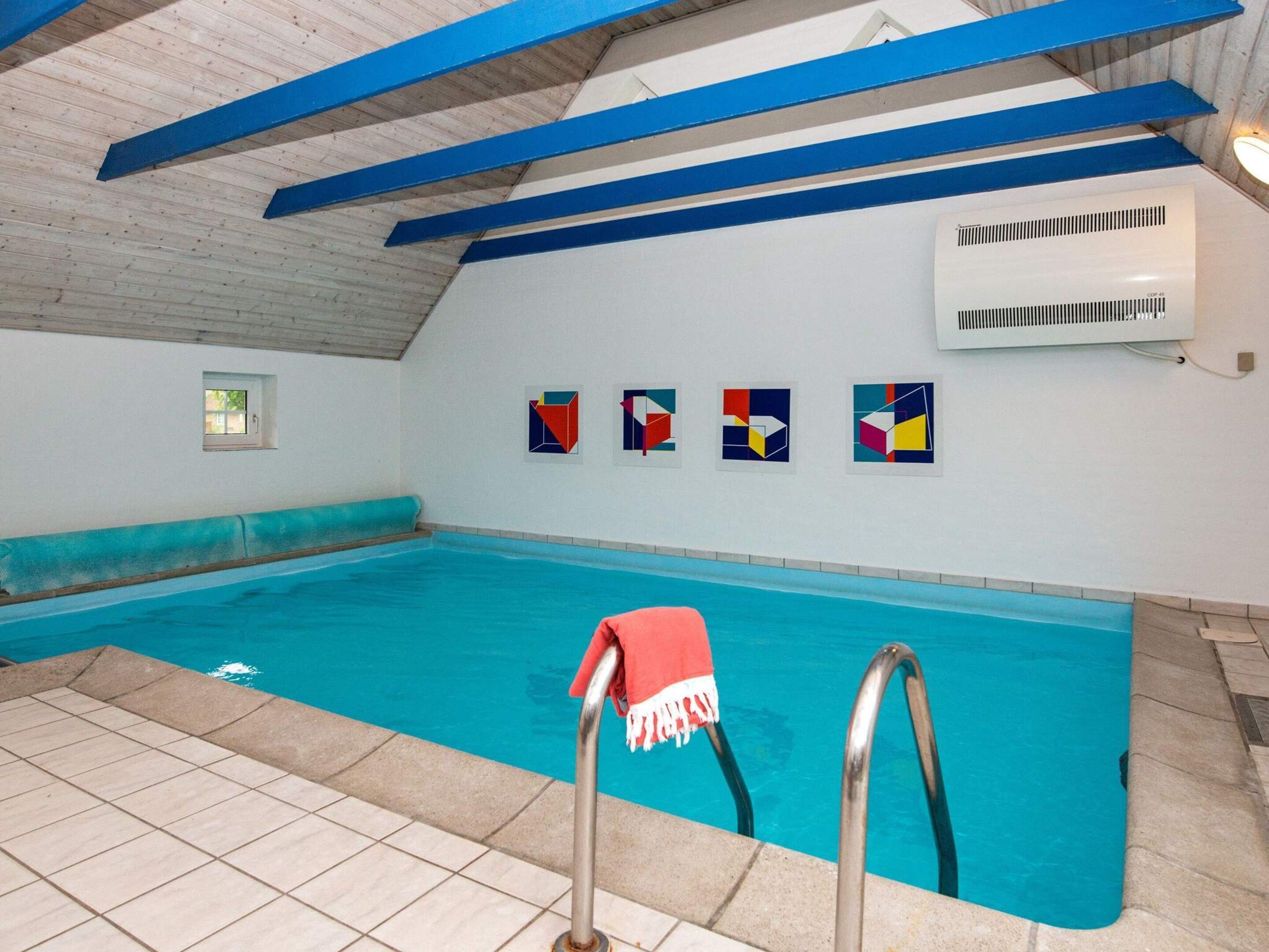 Una piscina interior con vigas azules, suelo de baldosas y una toalla roja en el escalón.