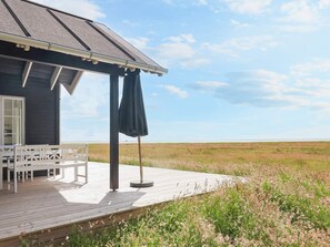 A wooden deck with a table and chairs, a covered area with a dark umbrella, and a view of a grassy field. - 6 Person Holiday Home in Laeso (Læsø)