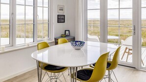 Un coin repas avec une table blanche et des chaises jaunes, de grandes fenêtres donnant sur une prairie, et un tableau encadré accroché au mur.