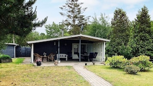 Einbungalow mit überdachter Veranda, einer Terrasse mit Gartenmöbeln und einer gepflasterten Auffahrt zum Eingang.