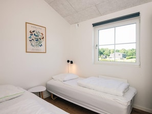 Une chambre avec un lit simple, une petite table de chevet, une estampe botanique encadrée accrochée au mur et une fenêtre donnant sur un immeuble et des arbres.