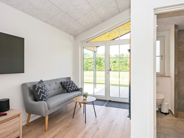 Ein modernes Wohnzimmer mit einem grauen Sofa, zwei gemusterten Kissen, einem kleinen runden Tisch mit einer Pflanze und einem großen Fenster mit Blick auf Grünflächen.