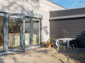 Un bâtiment moderne avec une entrée dotée d’une porte en verre, un patio aménagé avec une table et des chaises, et des plantes en pot.
