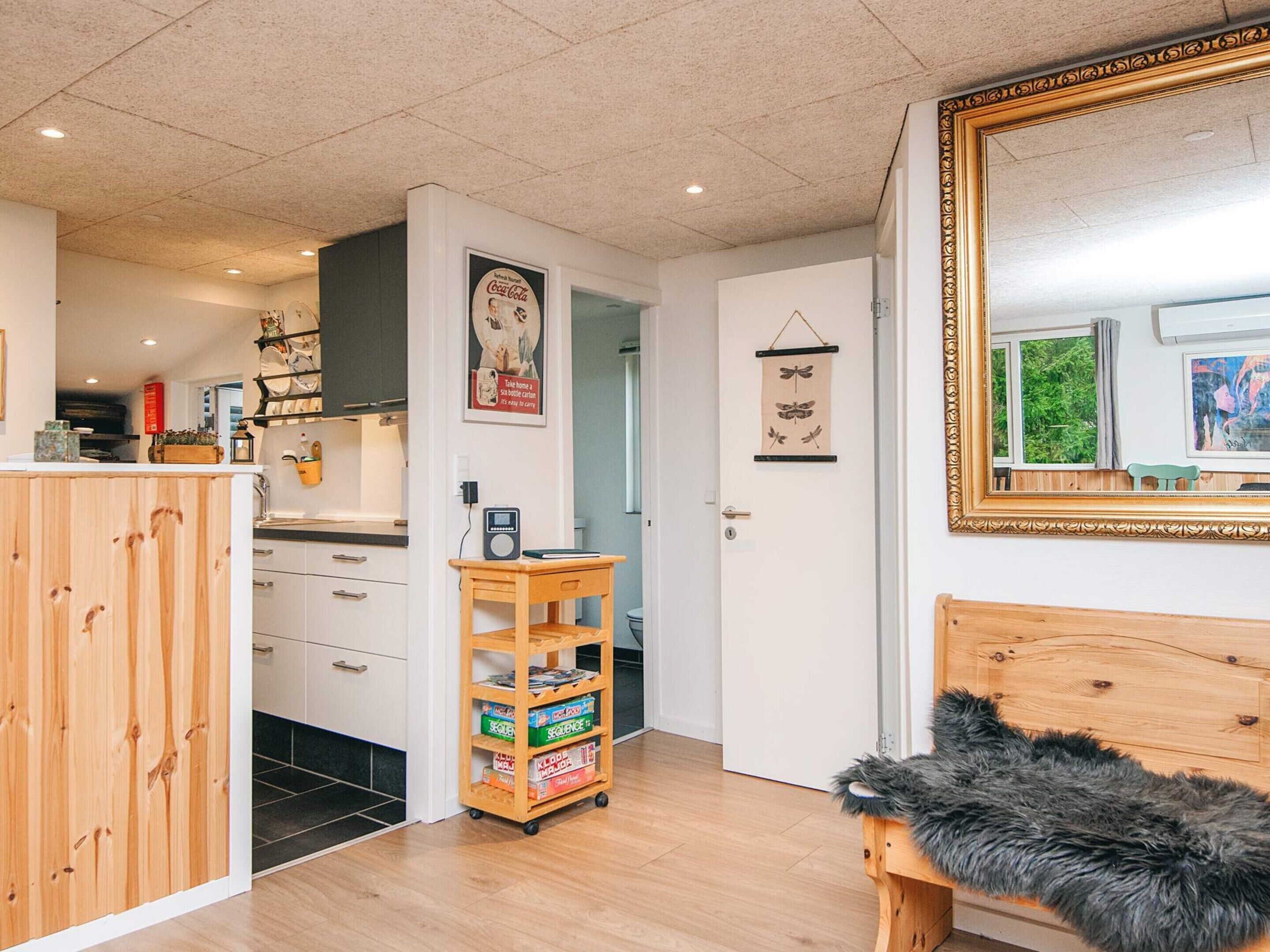 A compact living space with a wooden cabinet, a small wooden table with a box of board games, a bench with a fur throw, a framed mirror, and a door with a hanging wall decoration.