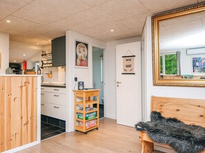 A compact living space with a wooden cabinet, a small wooden table with a box of board games, a bench with a fur throw, a framed mirror, and a door with a hanging wall decoration. - 8 Person Holiday Home in Ebeltoft (Ebeltoft)
