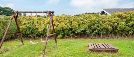 A wooden swing set in a grassy yard with a hedge and a house in the background.