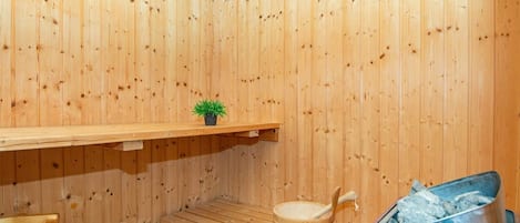 Un sauna de madera con un banco, un pequeño estante con una planta, un balde de madera y un recipiente rojo.
