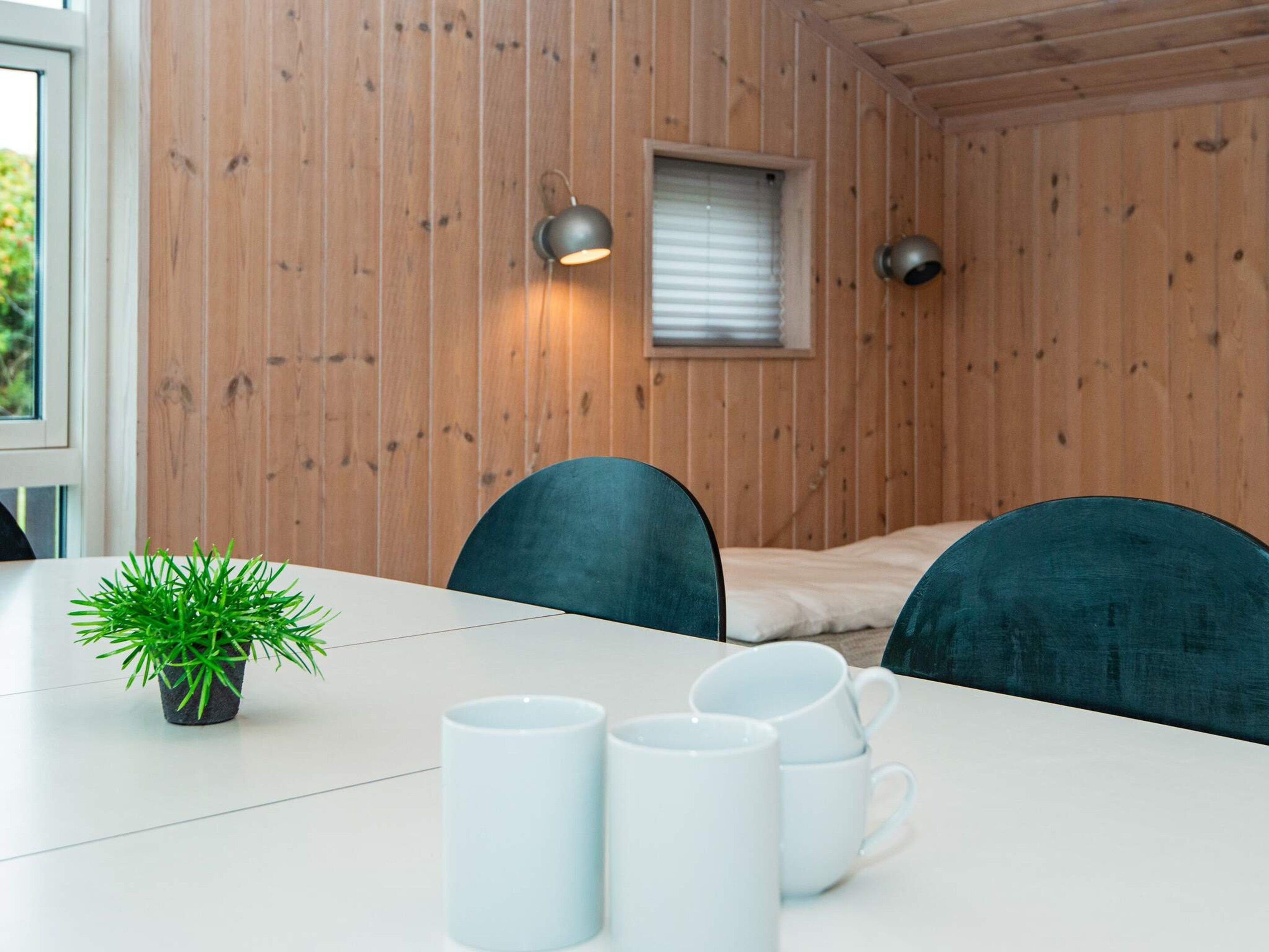 A room with a wooden wall, a bed, a window with blinds, a table with chairs, and a small potted plant.