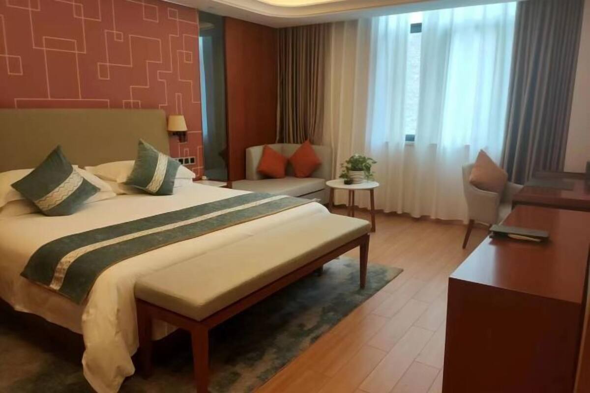 A hotel room with a bed, a desk, a chair, a small table with a plant, and a view of the outdoors through curtains.