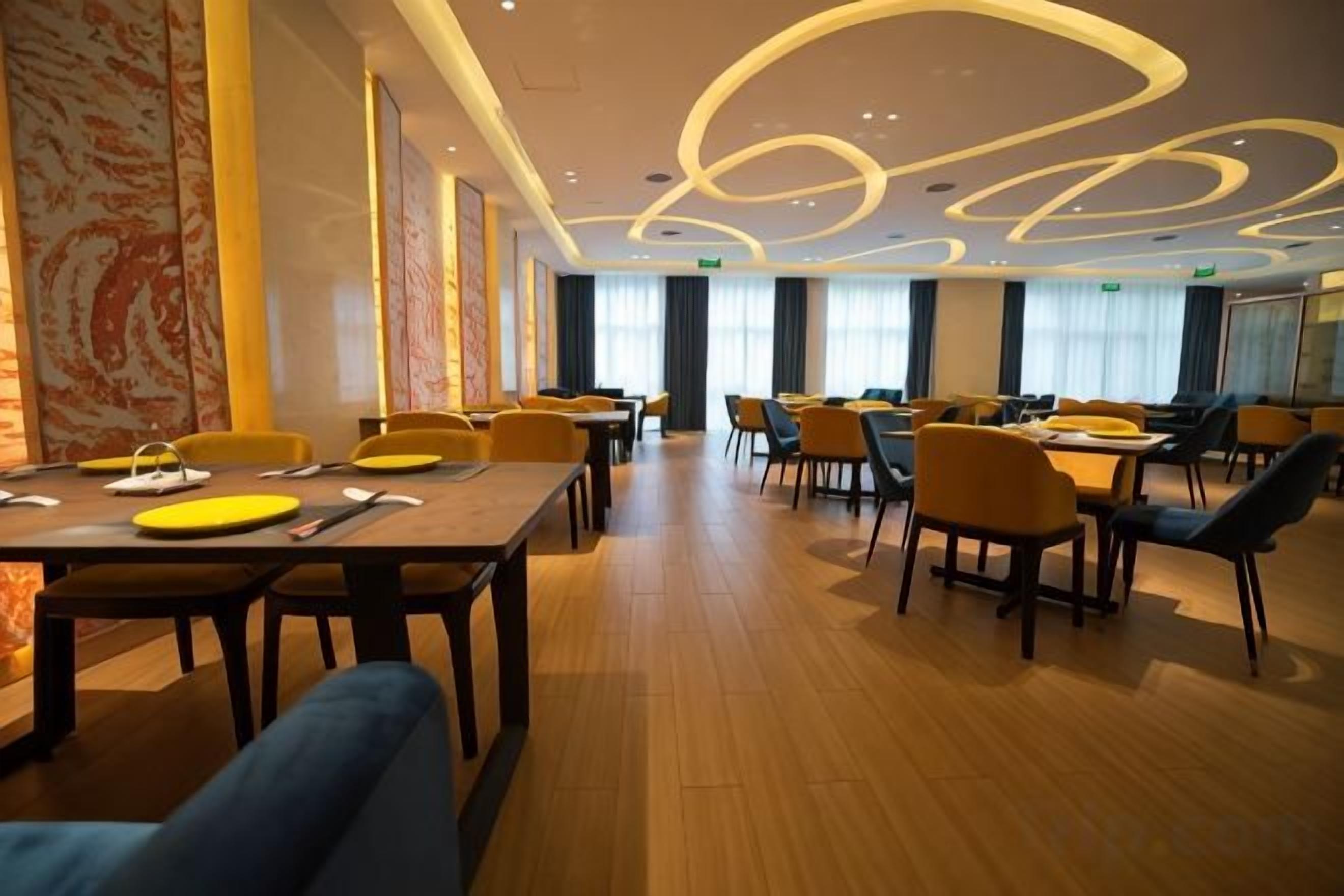 Een restaurant in een hotel, met meerdere tafels en stoelen, een modern plafond en grote ramen.