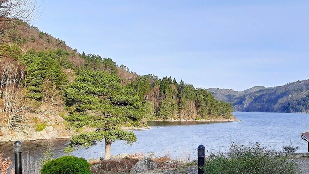 A wooden deck overlooking a lake with trees and hills in the background. - 9 Person Holiday Home in Lyngdal (Lyngdal)