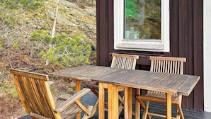 A wooden deck with a table and chairs, surrounded by a dark wooden building and a hillside with sparse vegetation. - 5 Person Holiday Home in Tyreso (Tyreso)