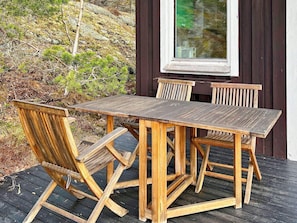 A wooden deck with a table and chairs, surrounded by a dark wooden building and a hillside with sparse vegetation. - 5 Person Holiday Home in Tyreso (Tyreso)