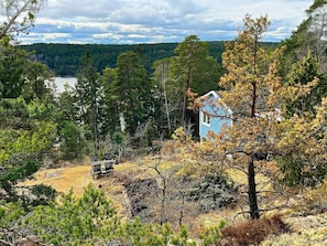 A cabin with a blue exterior, surrounded by dense forest and a lake in the background. - 5 Person Holiday Home in Tyreso (Tyreso)