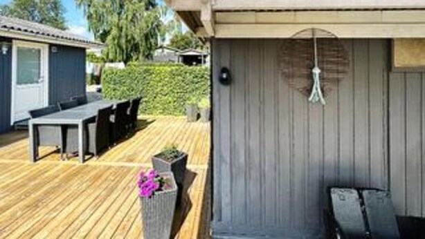 Una terraza de madera con mobiliario exterior, una maceta con flores y una lámpara colgante.