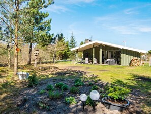 Une maison moderne dotée d’un porche abrité, d’un jardin avec des plantes et d’une mangeoire à oiseaux, ainsi que d’une pile de bois de chauffage.