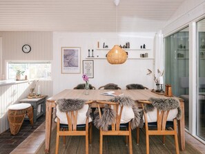 Un coin repas avec une table en bois, six chaises agrémentées de coussins en fausse fourrure, un luminaire suspendu et des étagères murales ornées d’objets décoratifs.