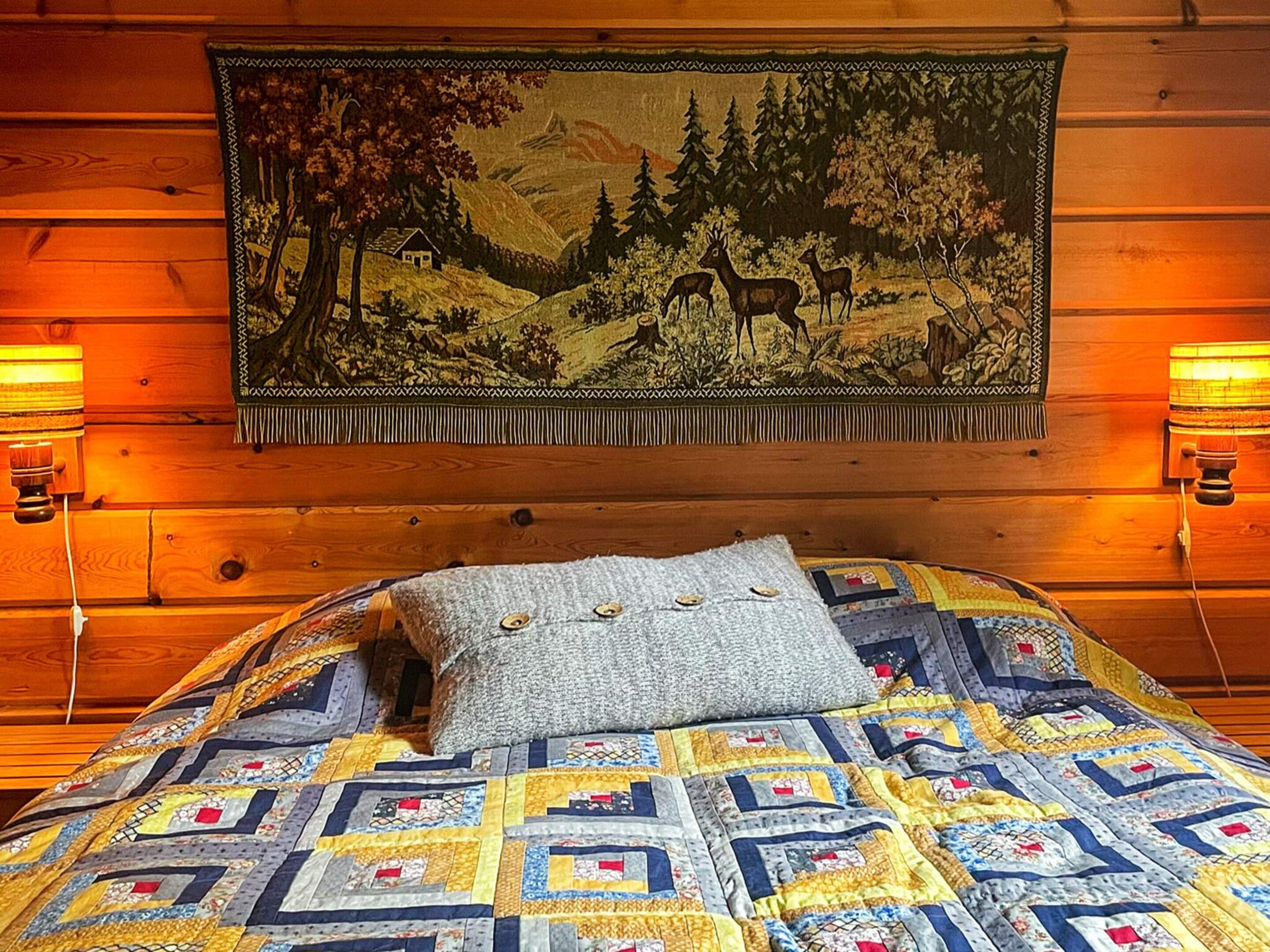 A bed with a patterned quilt and a grey pillow, a tapestry depicting a forest scene, and two wall-mounted lamps on either side of the bed.