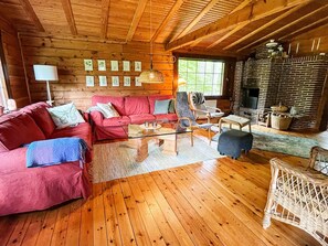 A cozy living room with a wooden ceiling, a brick fireplace, a red sofa, and a wicker chair.