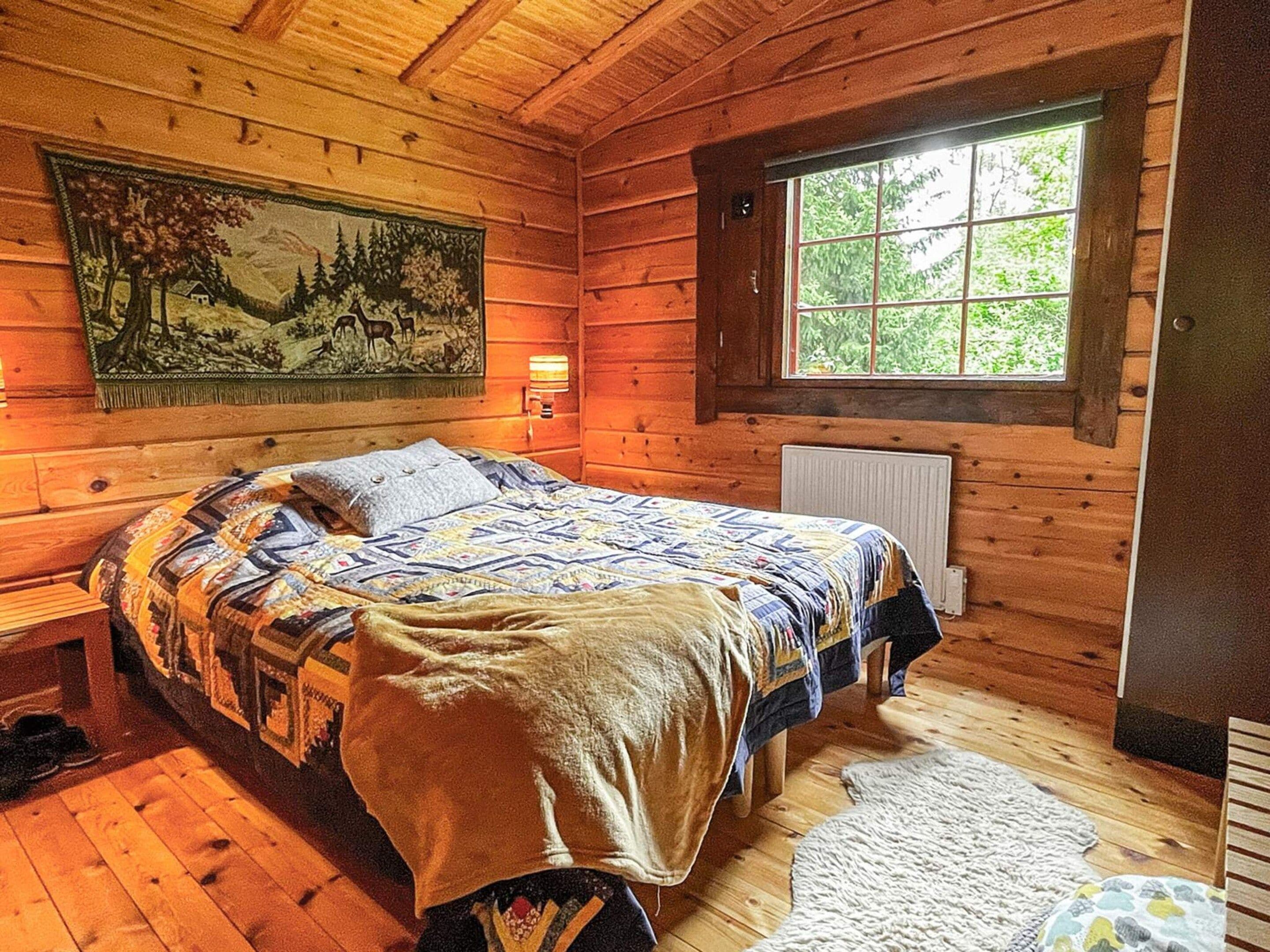A wooden cabin bedroom with a bed, a window, a radiator, and a painting on the wall.