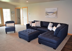 A living room with a sectional sofa, a chair, and an ottoman. There is a painting on the wall and a door in the background.