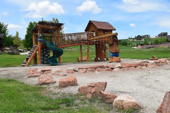 A wooden play structure with a slide and climbing area, surrounded by rocks and grass.