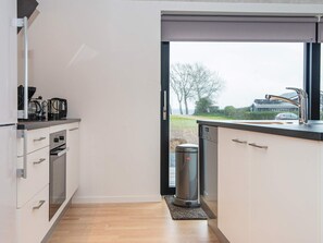 A modern kitchen with a built-in dishwasher, a coffee maker, and a view of a residential area through a large window. - 8 Person Holiday Home in Aabenraa (Aabenraa)