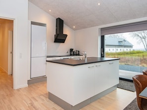 A modern kitchen with a central island, white cabinetry, a black range hood, and a view of a house through a large window. - 8 Person Holiday Home in Aabenraa (Aabenraa)