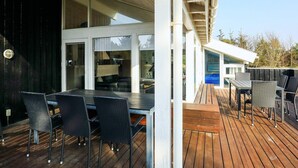 Una terraza exterior con suelo de madera, una mesa de comedor con sillas y un área de estar cubierta con vista al interior.