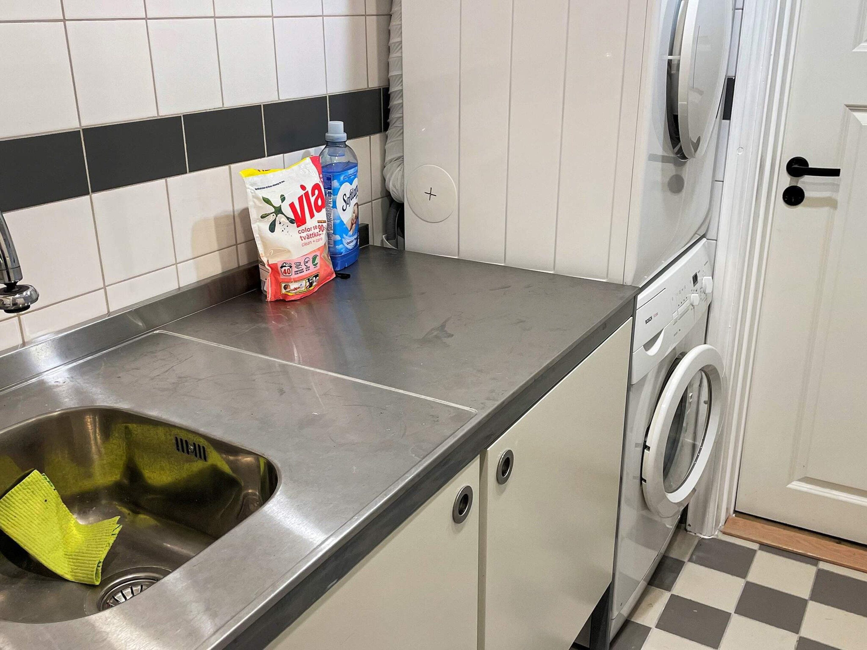 A kitchen with a stainless steel sink, a washing machine, and cleaning supplies on the counter.