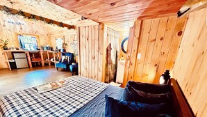A cozy cabin interior with a bed, a small kitchenette, a blue chair, and a wooden ceiling.