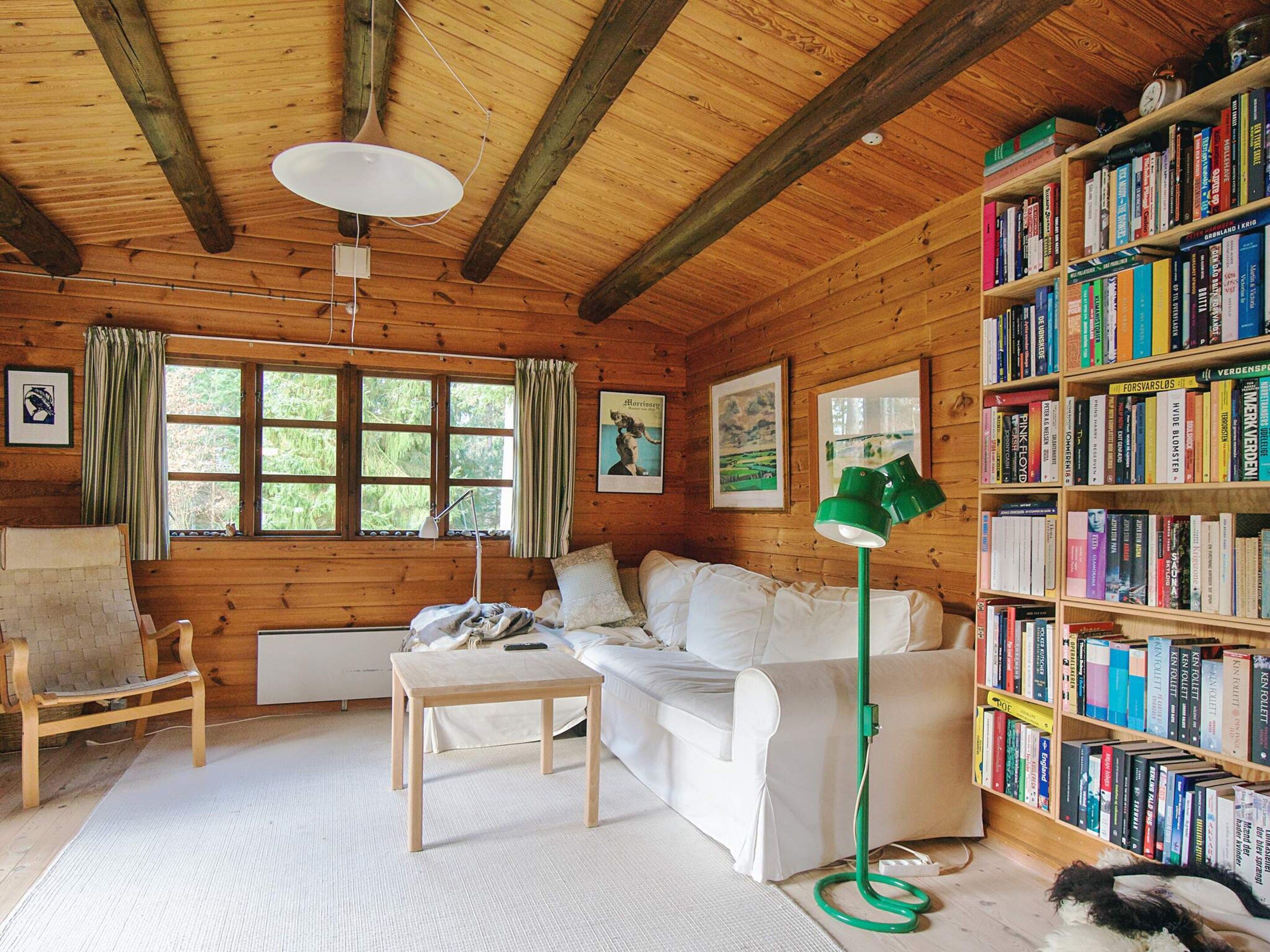 Una cabaña acogedora con interior de madera, un sofá blanco, una mesita, una estantería llena de libros y una ventana con cortinas.