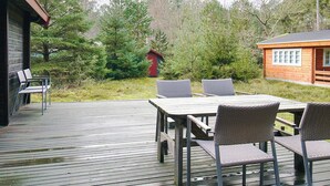 Une terrasse en bois avec une table et des chaises, entourée d’arbres et d’un petit abri de jardin rouge.